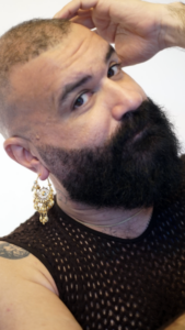 Photo of Khaled. He has a shaved head, black beard, gold dangly earring and black mesh vest
